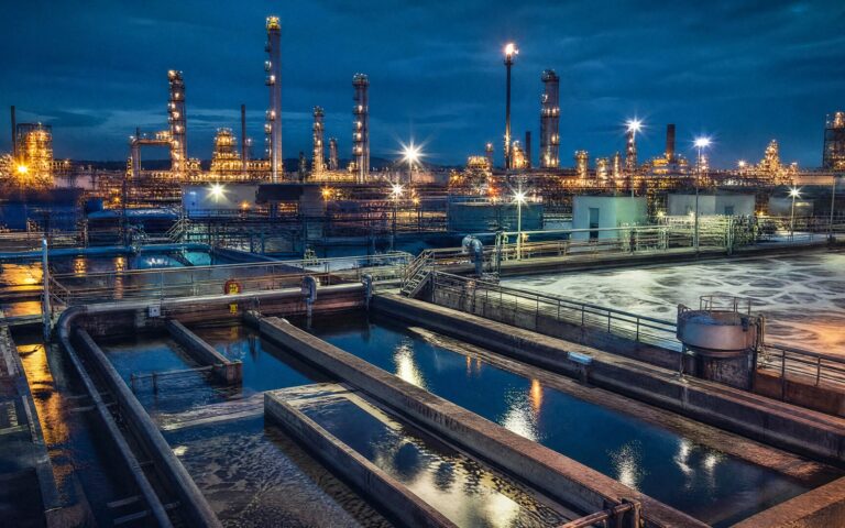 Gece aydınlatmalı rafineri arka planında arıtma havuzları ve boru hatlarıyla petrol rafinerisi atık su arıtma tesisi